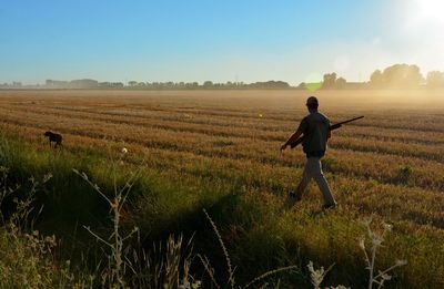 Rear view of man walking with rifle in farm against sky