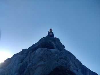 Low angle view of man sitting on cliff against clear blue sky