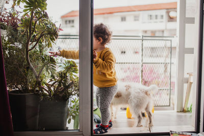 Full length of girl touching plants by window and dog