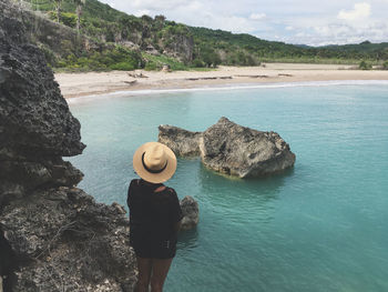 Rear view of woman wearing hat looking at sea while standing on rock