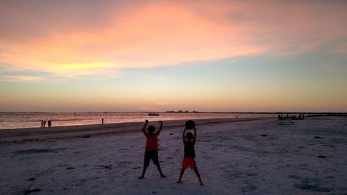 Full length of brothers standing at beach against sky during sunset