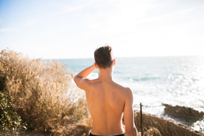 Rear view of shirtless teenage boy looking at sea against sky