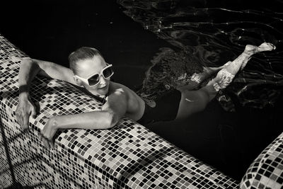 A vintage photo of a woman in a black swimsuit and white sunglasses swimming in the pool