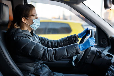 Woman with glasses, medical mask and latex gloves driving a car with her hands on steering wheel.