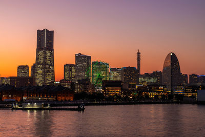 Illuminated buildings by river against sky during sunset