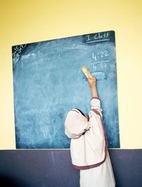 Rear view of girl erasing blackboard with duster while standing in classroom