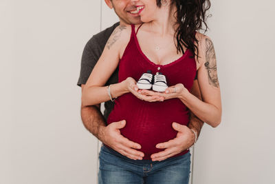 Midsection of pregnant couple holding shoes while standing against wall