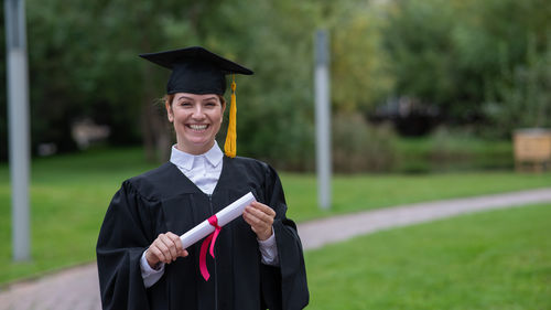 Portrait of smiling young woman wearing graduation gown standing on field