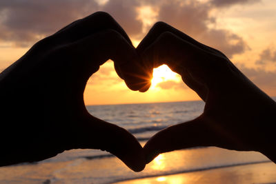 Cropped silhouette hands forming heart shape against sky during sunset