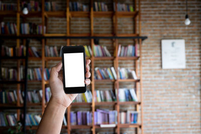 Cropped hand of woman holding mobile phone against bookshelf