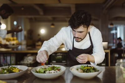 Focused male chef garnishing salad with cilantro while bending in commercial kitchen at restaurant