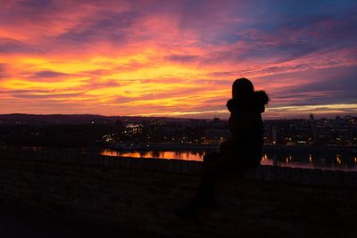 Silhouette woman sitting on retaining wall against orange sky