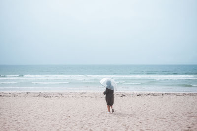 Back view of unrecognizable traveler in raincoat with transparent umbrella walking on empty sandy beach towards waving sea in cloudy day