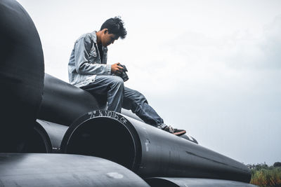 Low angle view of man looking at camera while sitting on pipes against sky