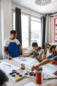 Smiling male teacher assisting pupil painting while sitting in art class at elementary school