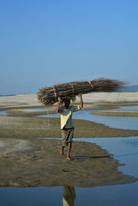 Man carrying logs at beach against clear blue sky