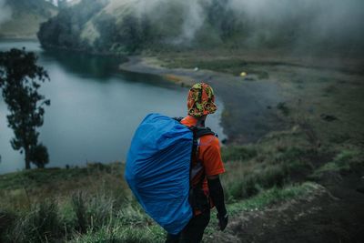 Rear view of hiker standing on field by lake during foggy weather