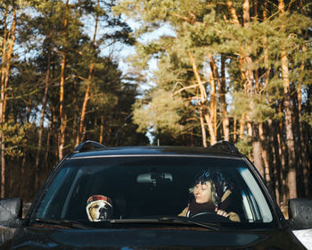 A woman and her dog wearing funny hats sit in the car at snowy forest. 