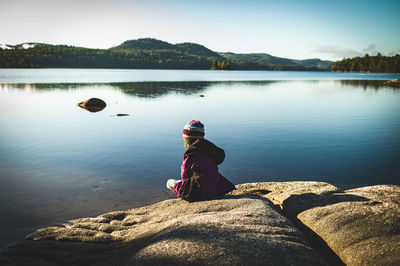 Girl sitting on rock by lake against sky