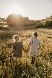 Rear view of male siblings holding hands and running in meadow at sunny day