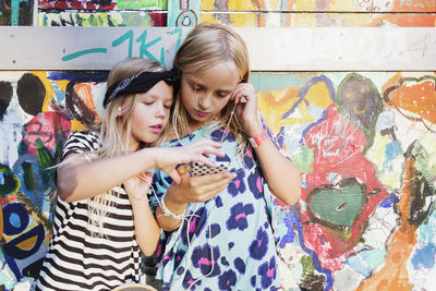 Girls listening music through mobile phone while standing against graffiti wall