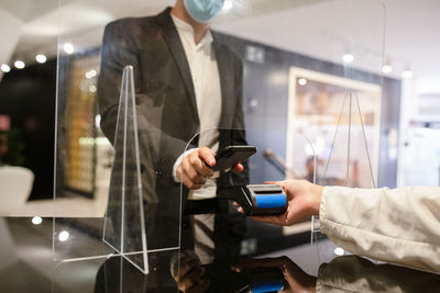 Crop anonymous male buyer in protective mask using smartphone to make contactless payment at reception desk with protective glass screen