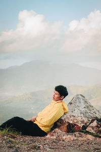 Side view of man sitting on mountain against cloudy sky