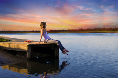 Side view of teenage girl sitting on pier over lake at sunset