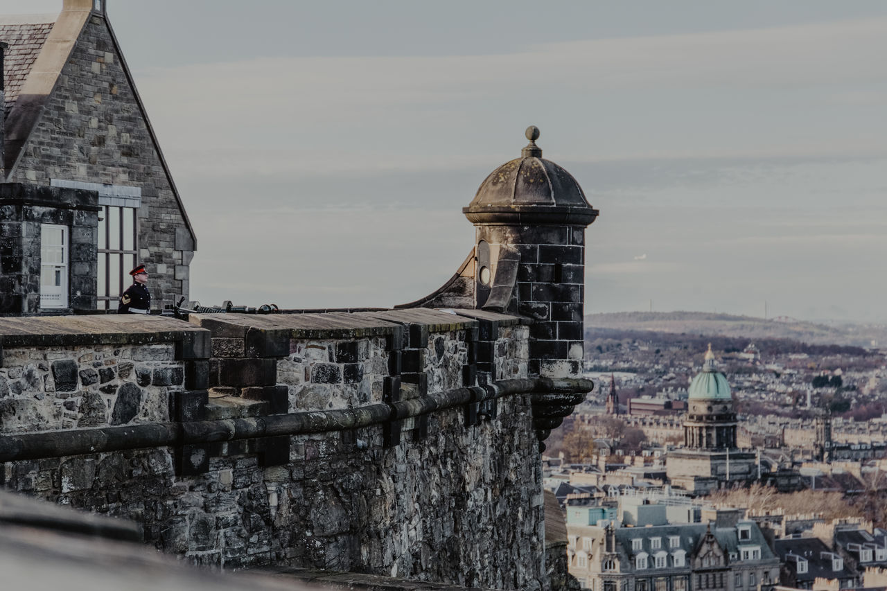 Male guard in uniform standing on roof of historic stone building against gray sky in city