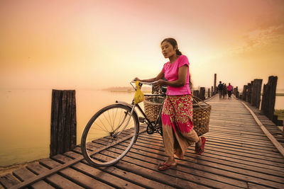 Woman riding bicycle by sea against sky during sunset
