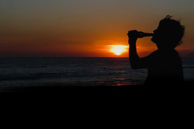 Silhouette man drinking while standing beach against orange sky during sunset