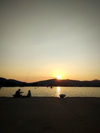 Silhouette people sitting on beach against sky during sunset