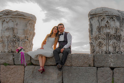 Portrait of smiling bride with groom sitting on stone wall at old ruin