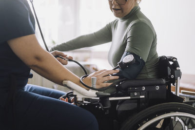 Midsection of female nurse checking blood pressure of woman sitting on wheelchair in clinic