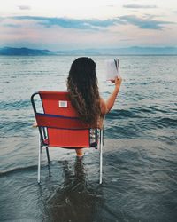 Rear view of woman sitting on chair while reading book in sea