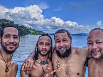Portrait of smiling shirtless friends at sea against sky
