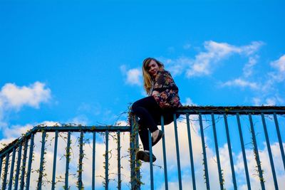 Low angle view of woman sitting on fence against blue sky