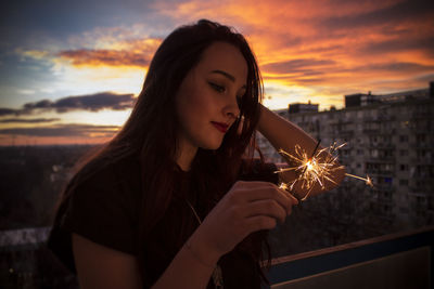 Close-up of young woman holding sparkler while standing against sky during sunset