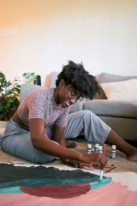 Young beautiful african american woman with afro hair, amateur artist in casual outfit drawing colorful picture with paintbrush on paper while sitting on floor in cozy apartment