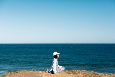 Side view of woman sitting on cliff by sea against clear sky