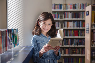 Portrait of smiling girl reading book while standing in library