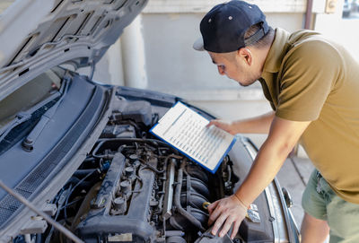 Side view of man holding checklist while looking at car engine