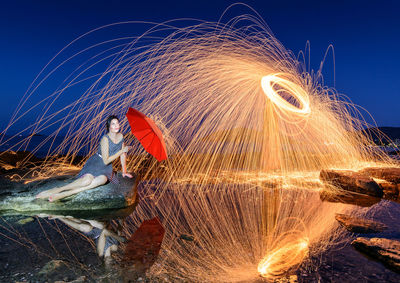 Blurred motion of woman standing by illuminated lights against sky at night