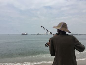 Rear view of man playing violin at beach against sky