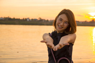 Portrait of smiling young woman gesturing while standing against sea and sky during sunset