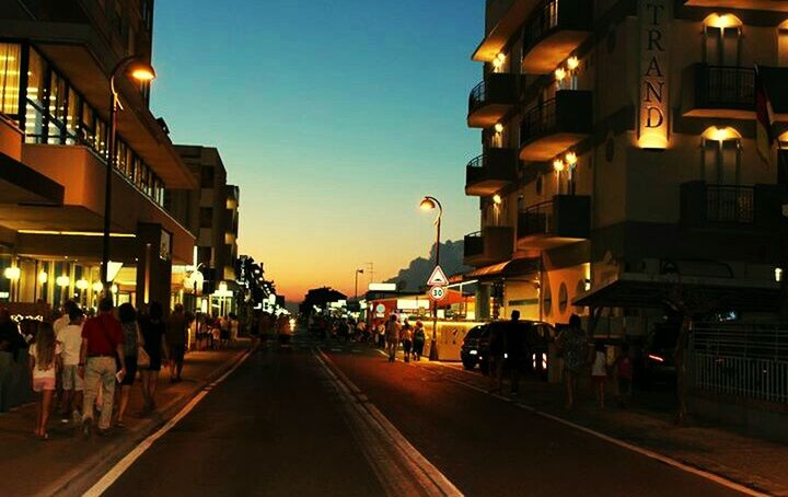 building exterior, architecture, illuminated, built structure, street, city, the way forward, diminishing perspective, city life, road, night, street light, dusk, incidental people, lighting equipment, transportation, sky, sunset, outdoors, clear sky