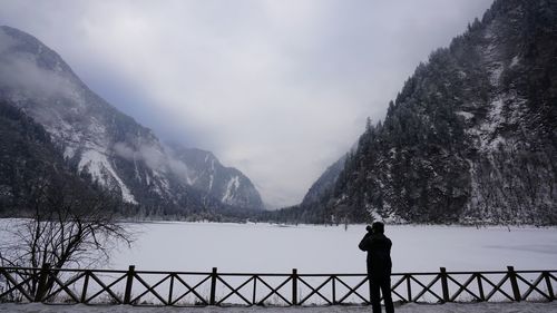 Man standing on railing by lake against sky during winter
