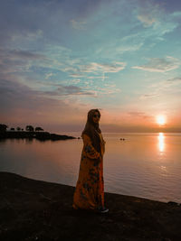 Woman standing by sea against sky during sunset