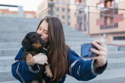 Beautiful young adult woman taking a selfie photo kissing her dog