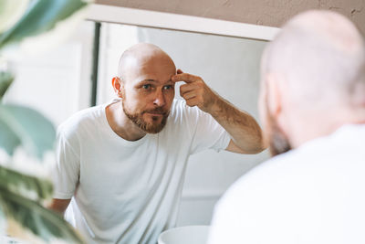 Handsome bald man looking at mirror and touching face in bathroom